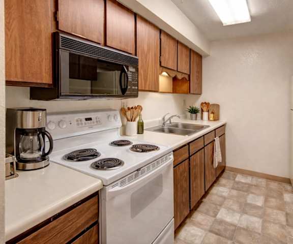 A cozy kitchen with white appliances, kitchen utensils, overhead lighting, light countertops with lots of room, and large wooden cabinets at Terrace on the Lake Apartment Homes. 