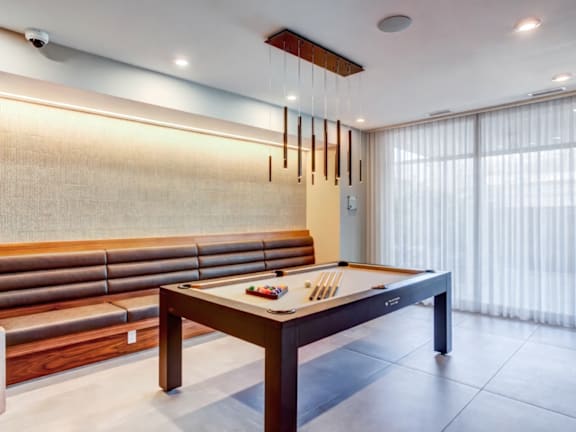 A pool table with cues and balls is in the middle of a room with a bench and curtains.