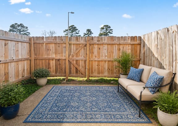 A blue rug sits on a wooden deck with a white couch and potted plants.