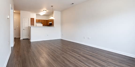 A room with a ceiling fan and wooden flooring with a kitchen in the background