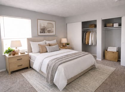 A bedroom with a large bed, a nightstand with a lamp, and a wardrobe with clothes hanging.