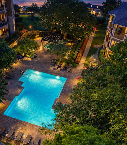A beautifully lit swimming pool surrounded by a well-manicured garden and residential buildings.