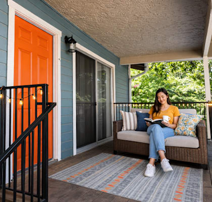 A woman is sitting on a couch on a porch with a book in her hands.