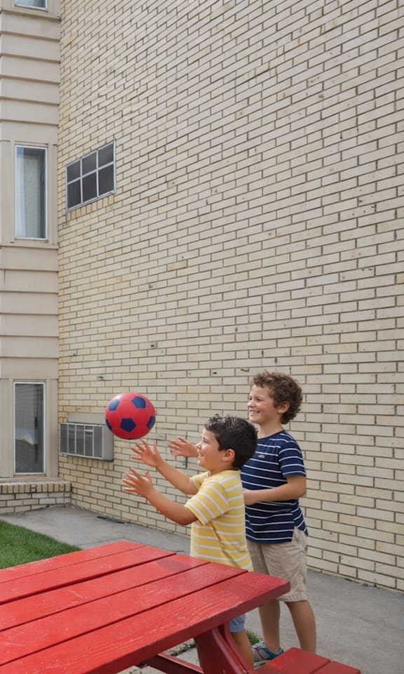 A man and a woman are standing by a grill with two children playing with a ball.