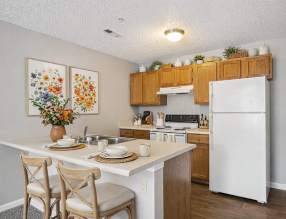 A kitchen with a white fridge, wooden cabinets and a table with two chairs.