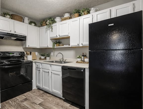 A black fridge stands in a kitchen with white cabinets and a wooden floor.