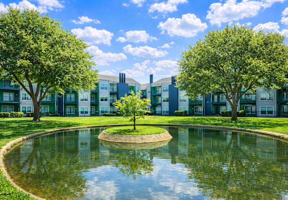 A small pond in the middle of a grassy area with trees and apartment buildings in the background. at Infinity on the Landing Apartments , Apartments, Euless, TX