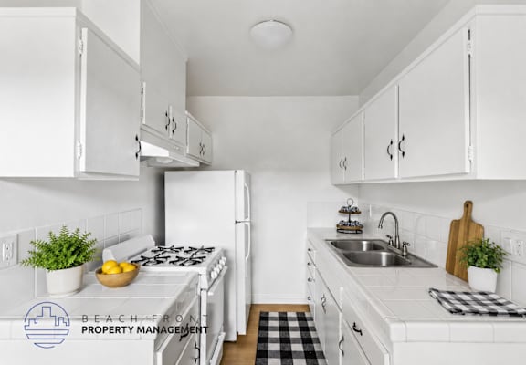 A kitchen with white cabinets and appliances, a black and white checkered mat on the floor, and a logo for Beach Front Property Management in the corner.