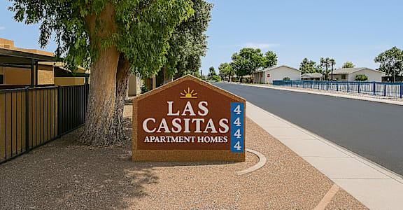 A brown sign with the words "Las Casitas Apartment Homes" in front of a tree.