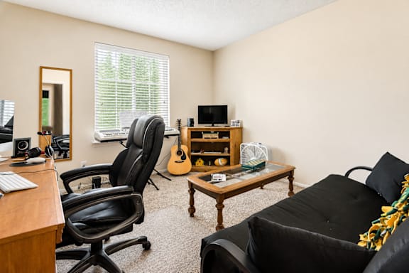 A black chair is in front of a desk with a computer and a guitar in the background.