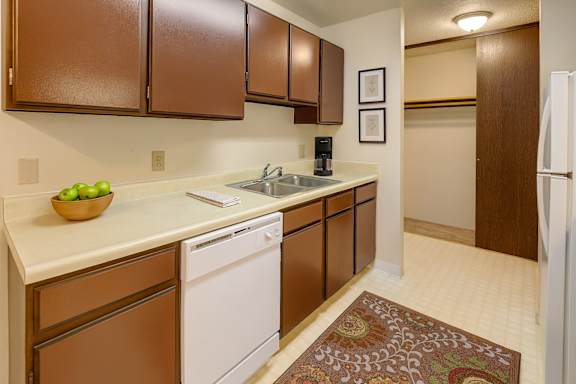 A spacious/cozy kitchen with white appliances, kitchen utensils, overhead lighting, light countertops with lots of room, utensils, and large medium wooden cabinets at Taiga Apartment Homes.