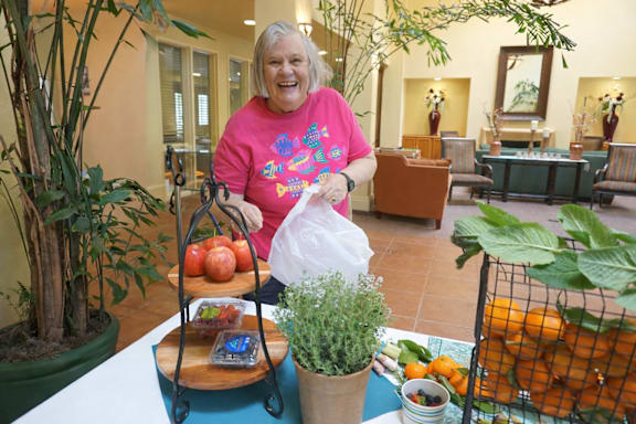 A woman in a pink shirt is holding a plastic bag and standing in front of a table with a tray of fruit and a potted plant.