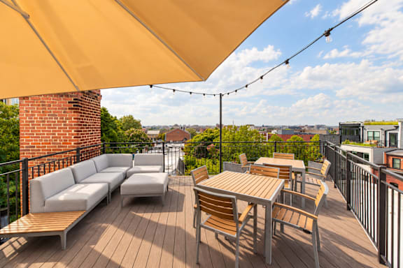 A patio with a white couch and wooden chairs under a yellow umbrella.