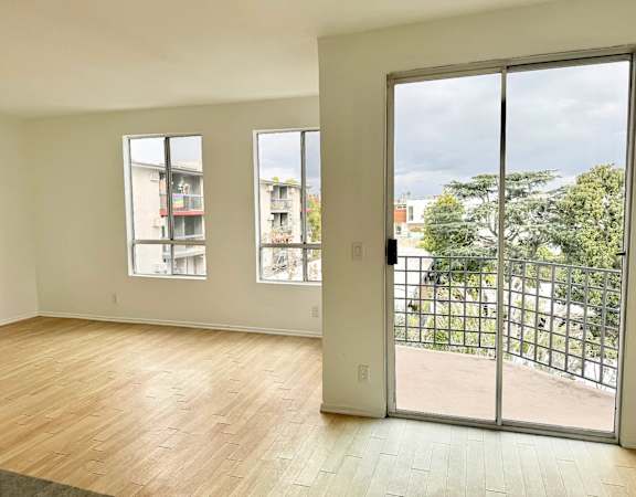 A room with a sliding glass door leading to a balcony with a view of a tree and a building.
