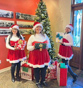 Three people dressed as Santa Claus stand in front of a Christmas tree.