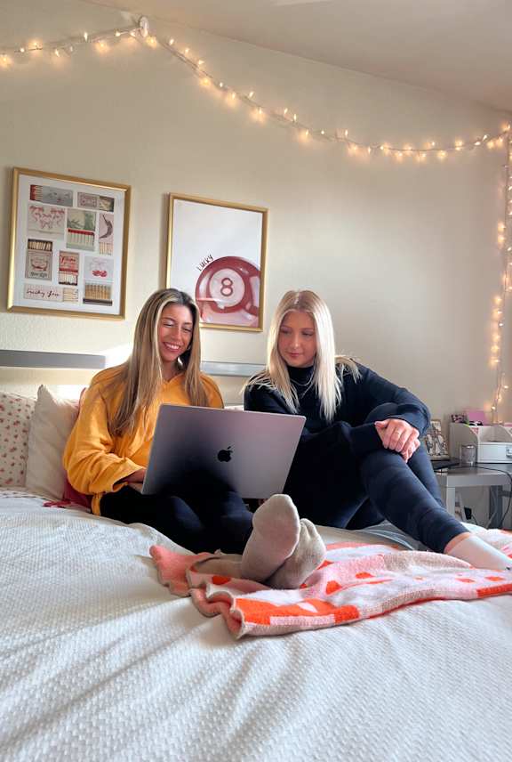 Two women sitting on a bed with laptops.