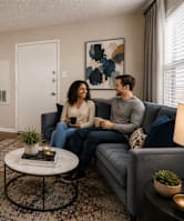A man and woman are sitting on a blue couch in a living room with a small tree, a coffee table, and a lamp.