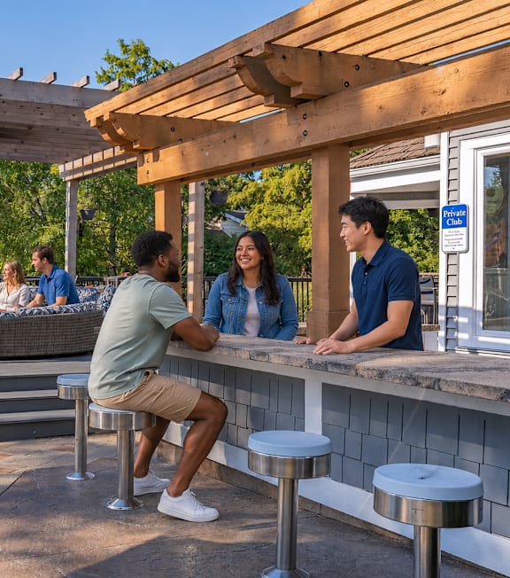 A group of people are sitting and standing around a picnic table under a wooden pergola.