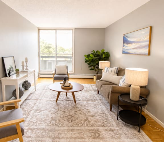 A living room with a grey rug, a grey sofa, a wooden chair, a wooden coffee table, a white side table, a white wall, a window, a plant, and a painting.