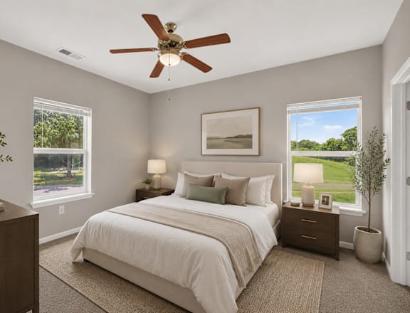 A bedroom with a large bed, a ceiling fan, and a view of a green landscape through the windows.