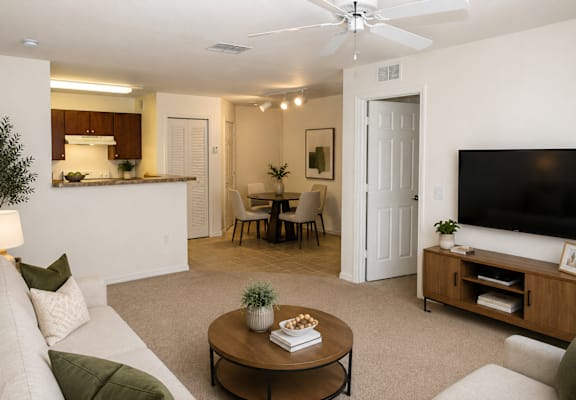 A living room with a white couch, a coffee table, a television, and a dining area in the background.