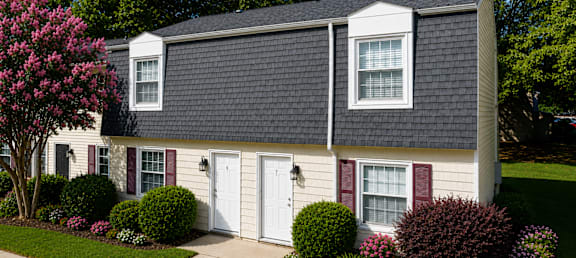 A house with a white door and red window frames is surrounded by greenery and a tree with pink flowers.