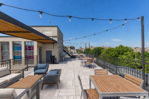A patio with wooden tables and chairs is covered with a roof and has string lights hanging above it.