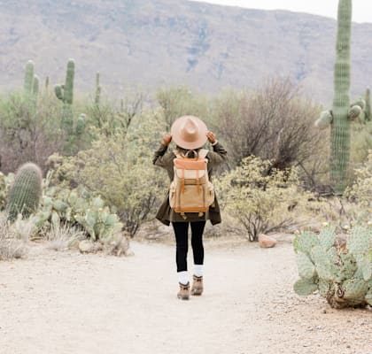 a woman with a backpack walking on a dirt road with cactus and mountains