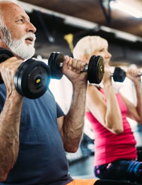 An older man and a younger woman are working out with dumbbells in a gym.