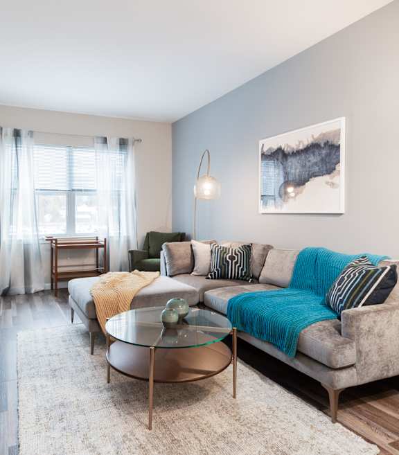 A living room with a grey couch, a glass table, and a painting on the wall.