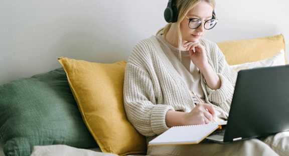 A person is sitting on a couch with a laptop and notebook, wearing headphones.