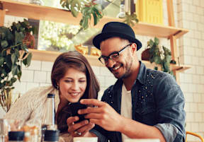 A man and a woman are sitting at a table in a cafe, with the man looking at a smartphone.