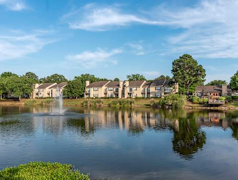 A serene lake with a fountain in the middle, surrounded by houses and trees under a clear blue sky. at Reflection Pointe, Flowood, 39232