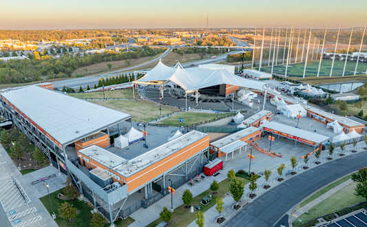 A large, modern building with a white and orange roof is surrounded by a parking lot and a grassy area.