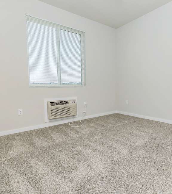A room with a carpeted floor, a window with blinds, and a wall-mounted air conditioning unit.