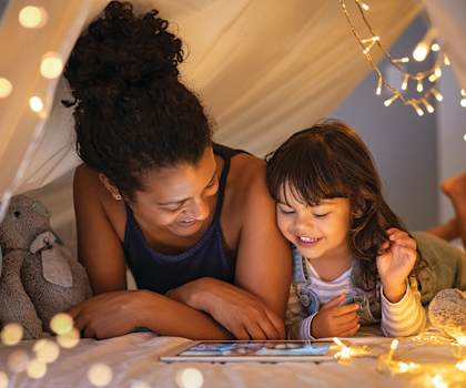 A woman and a child are lying in a tent-like bed with a tablet in front of them.
