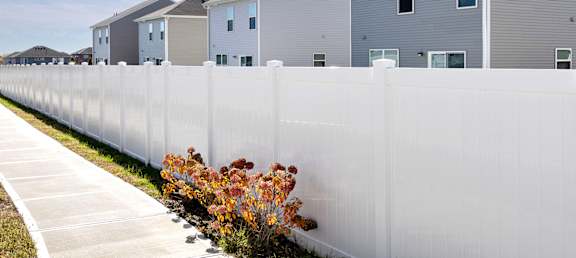 A white fence with a bush with orange leaves in front of a grey house.