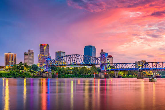 A city skyline at dusk with a bridge and buildings reflected in the water.