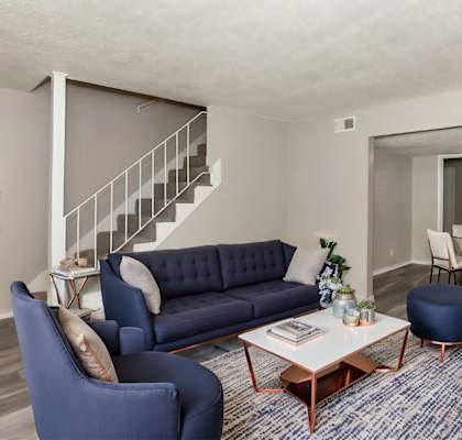 A living room with a blue sofa, a blue armchair, a wooden coffee table, and a staircase in the background.