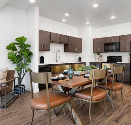 Modern apartment kitchen and dining area with wooden table, brown chairs, dark cabinets, and black appliances.