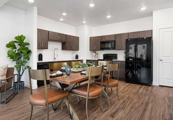 Modern apartment kitchen and dining area with wooden table, brown chairs, dark cabinets, and black appliances.