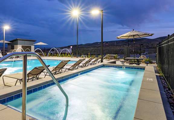 Outdoor swimming pool and spa area with lounge chairs, umbrellas, and evening lighting overlooking scenic hills.