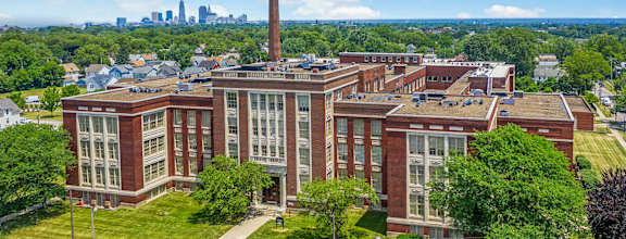 A large red brick building with a tall chimney is surrounded by trees and has a city skyline in the background.