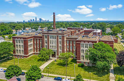 A large red brick building with a tall chimney is surrounded by trees and has a city skyline in the background.
