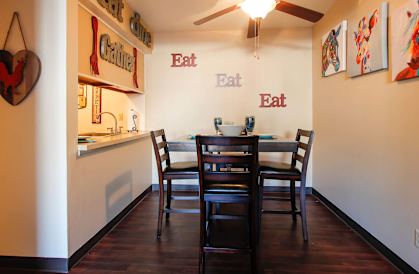 A dining room with a table and chairs and a wall hanging that says eat.