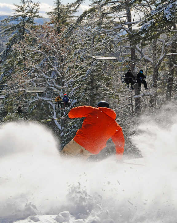 A skier in a red jacket is seen in the foreground of the image, with a large splash of snow behind them.