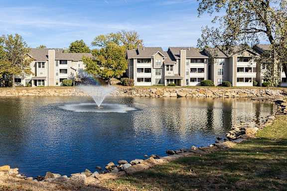 Scenic view of Three Lakes Apartments with a peaceful lake, a fountain, and apartment buildings surrounded by greenery.