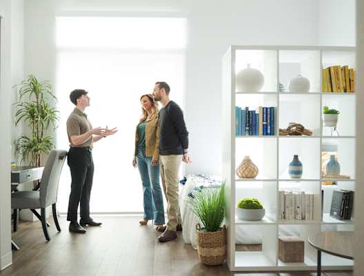 Three people are standing in a room with a bookshelf filled with books and decorative items.