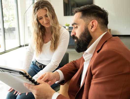 A man in a brown suit is holding a clipboard and appears to be explaining something to a woman in a white shirt who is sitting across from him.