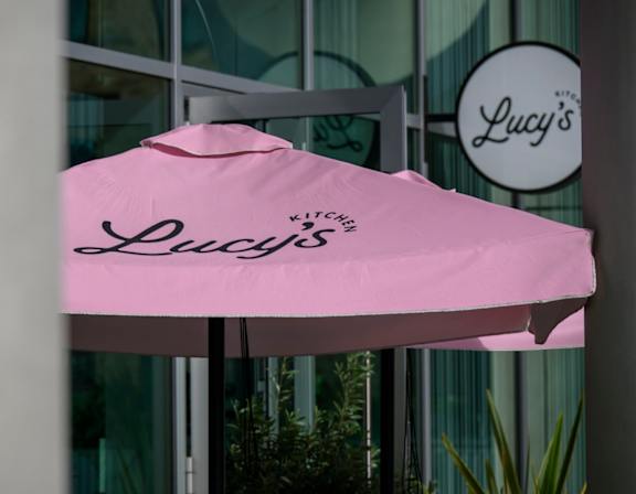 A pink umbrella with the word Lucy's on it is in front of a glass window.